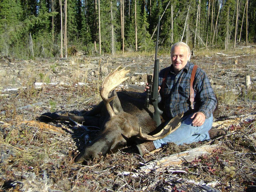 A moose hunter holding a rifle and sitting next to a newly-bagged moose